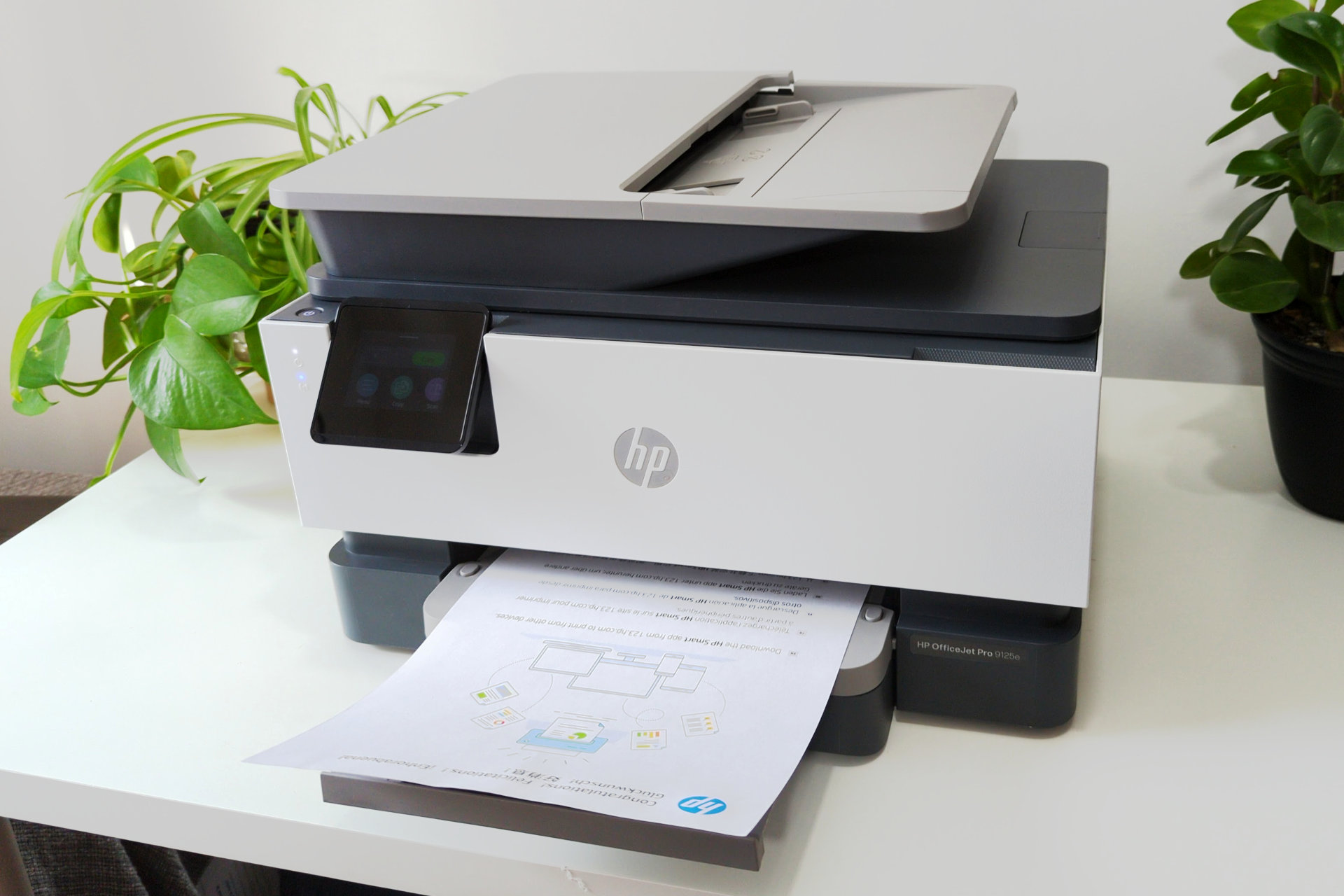 HP OfficeJet Pro 9125e روی یک پایه سفید رنگ که توسط گیاهان احاطه شده است قرار دارد.