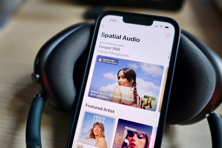An Apple iPhone 14 displaying the Apple Music app with a feature page on Spatial Audio content, beside a set of Apple AirPods Max headphones.