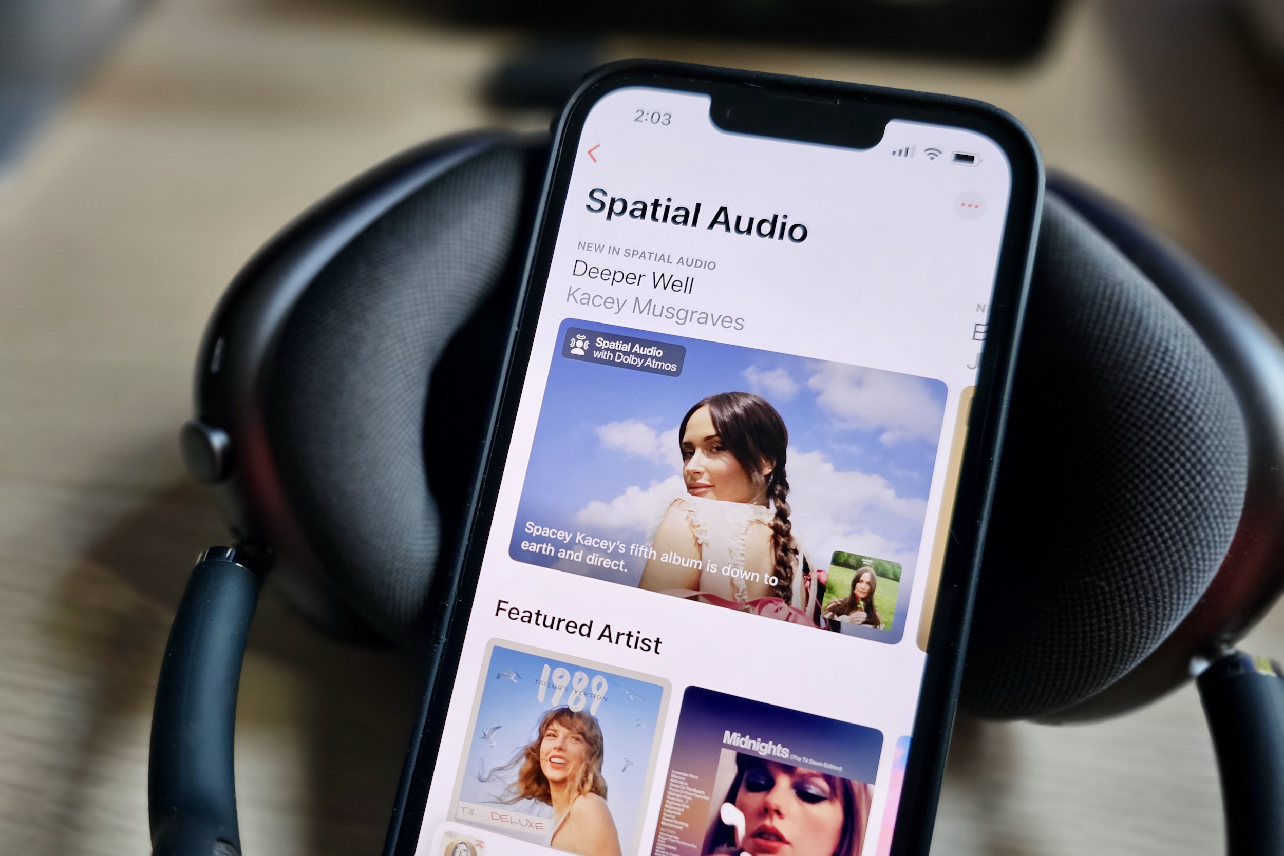 An Apple iPhone 14 displaying the Apple Music app with a feature page on Spatial Audio content, beside a set of Apple AirPods Max headphones.