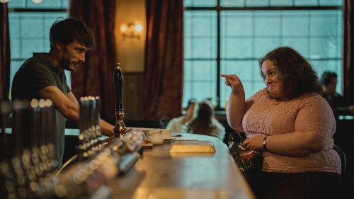 A man behind a bar, and a heavyset woman sitting there pointing at him, both smiling in a scene from Baby Reindeer.