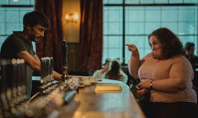 A man behind a bar, a heavyset woman sitting there and pointing at him, both smiling in a scene from Baby Reindeer.
