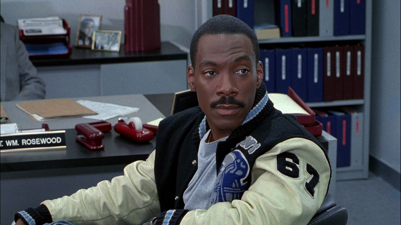 Eddie Murphy sentado com sua jaqueta icônica em um escritório na Beverly Hills Cop.