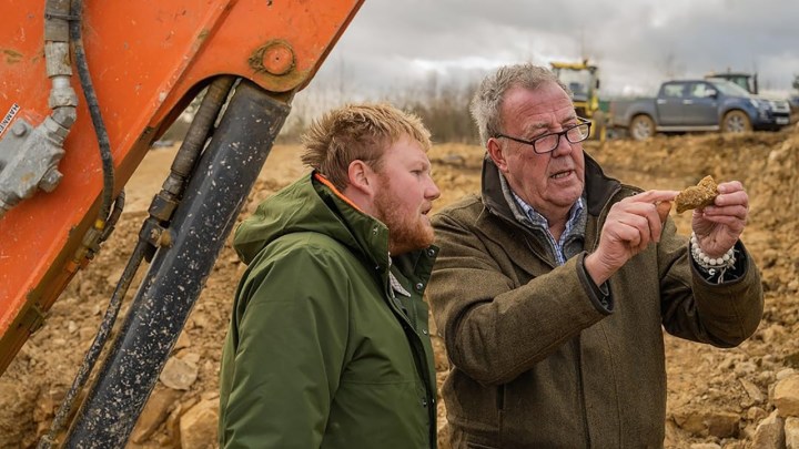 Two men standing in a farm looking at a piece of rock together by a tractor in a scene from Clarkson's Farm.