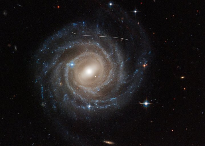 这张由 NASA/ESA 哈勃太空望远镜拍摄的棒旋星系 UGC 12158 的图像看起来像是有人用白色记号笔在上面画的。实际上，它是前景小行星穿过哈勃视野的时间曝光的组合，对银河系的观测进行了照片轰炸。对该星系进行了多次曝光，虚线图案证明了这一点。