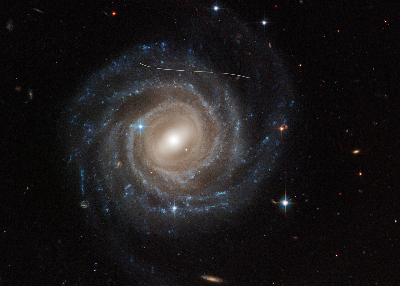 Это изображение спиральной галактики UGC 12158 с перемычкой, полученное космическим телескопом «Хаббл» НАСА/ЕКА, выглядит так, будто кто-то поднес к нему белую маркировочную ручку. На самом деле это комбинация временных экспозиций астероида на переднем плане, движущегося через поле зрения Хаббла, фотобомбящего наблюдение за галактикой. Было сделано несколько снимков галактики, о чем свидетельствует пунктирный рисунок.