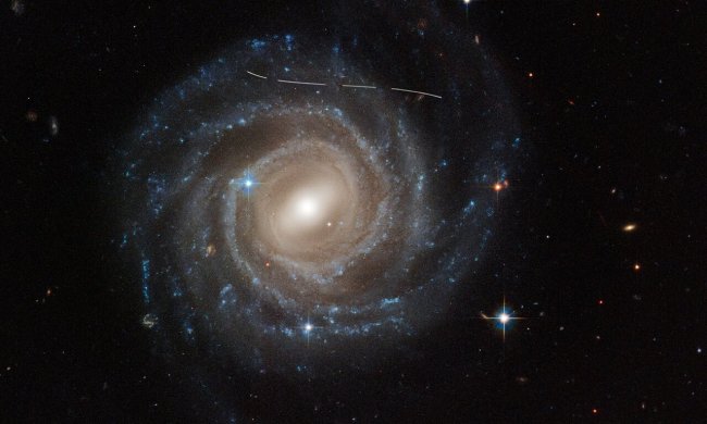 This NASA/ESA Hubble Space Telescope image of the barred spiral galaxy UGC 12158 looks like someone took a white marking pen to it. In reality it is a combination of time exposures of a foreground asteroid moving through Hubble’s field of view, photobombing the observation of the galaxy. Several exposures of the galaxy were taken, which is evidenced by the dashed pattern.