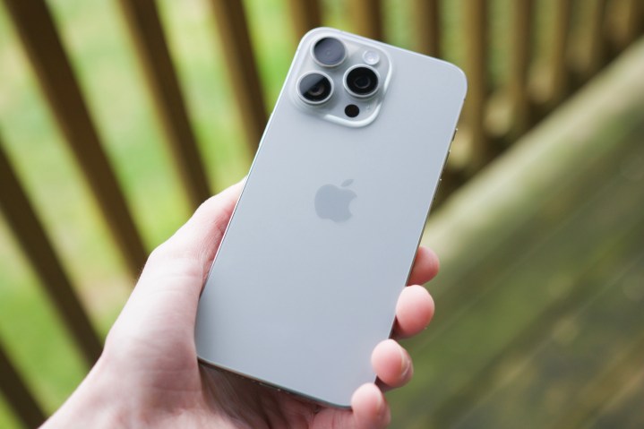 有人在户外露台上拿着 iPhone 15 Pro Max，展示了自然钛色的背面。