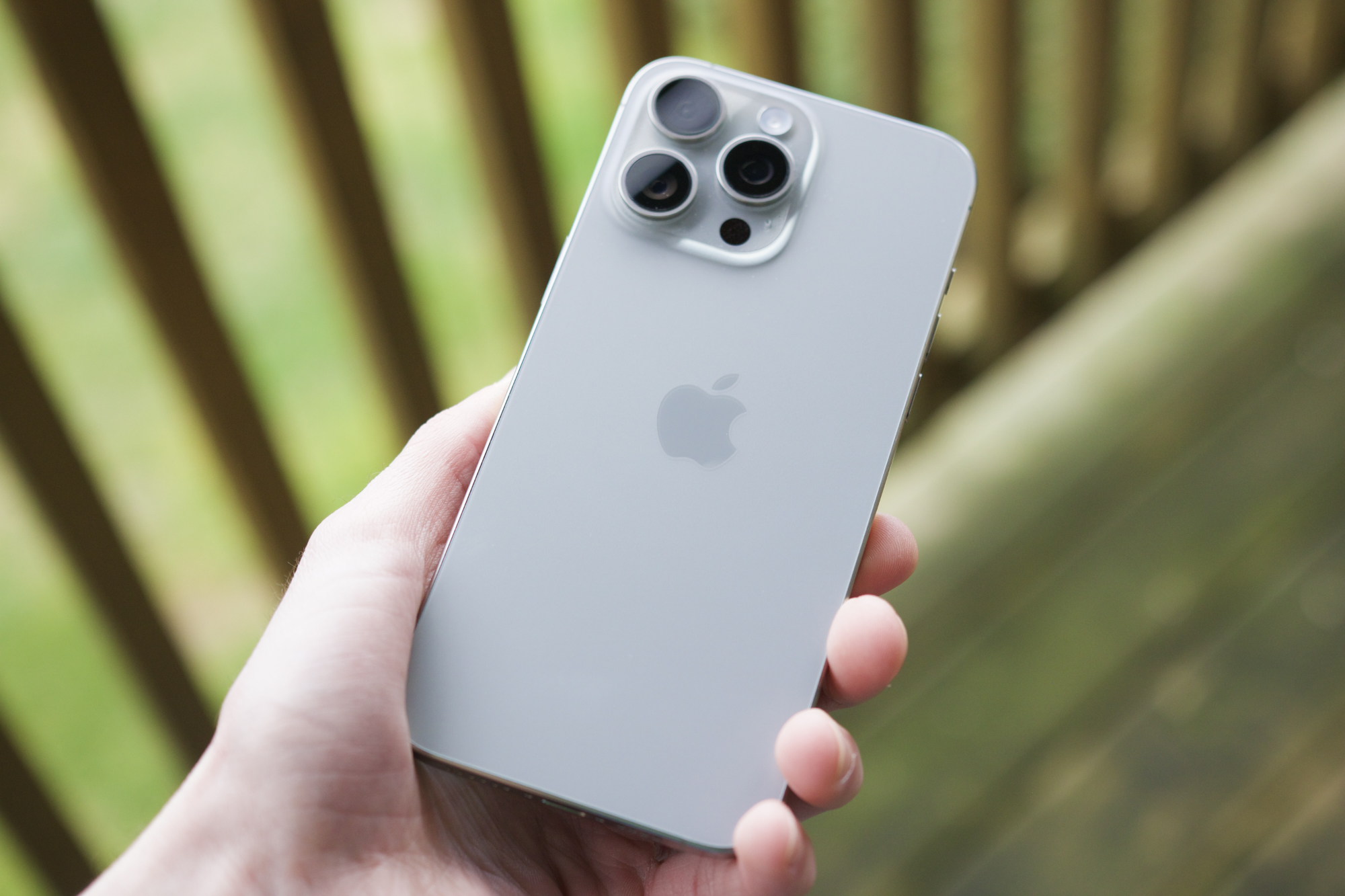 Alguém segurando um iPhone 15 Pro Max do lado de fora em um pátio, mostrando a parte traseira da cor Natural Titanium.