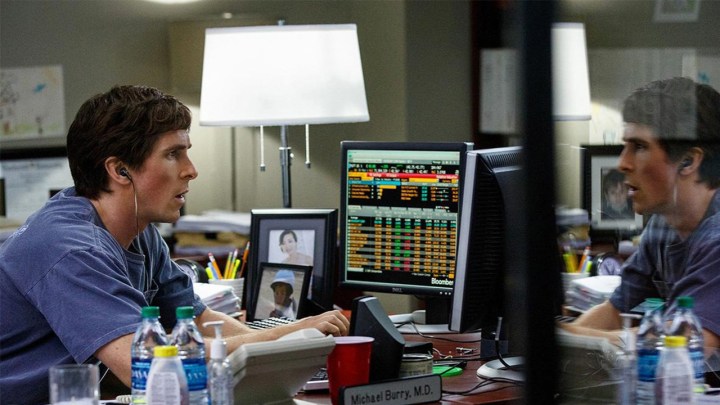 Christian Bale sentado en su escritorio mirando la pantalla de una computadora con información financiera, su reflejo visible frente a él en una escena de The Big Short.