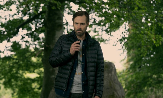 Will Forte standing outside in a black puffy jacket talking into a recorder in a scene from Bodkin.