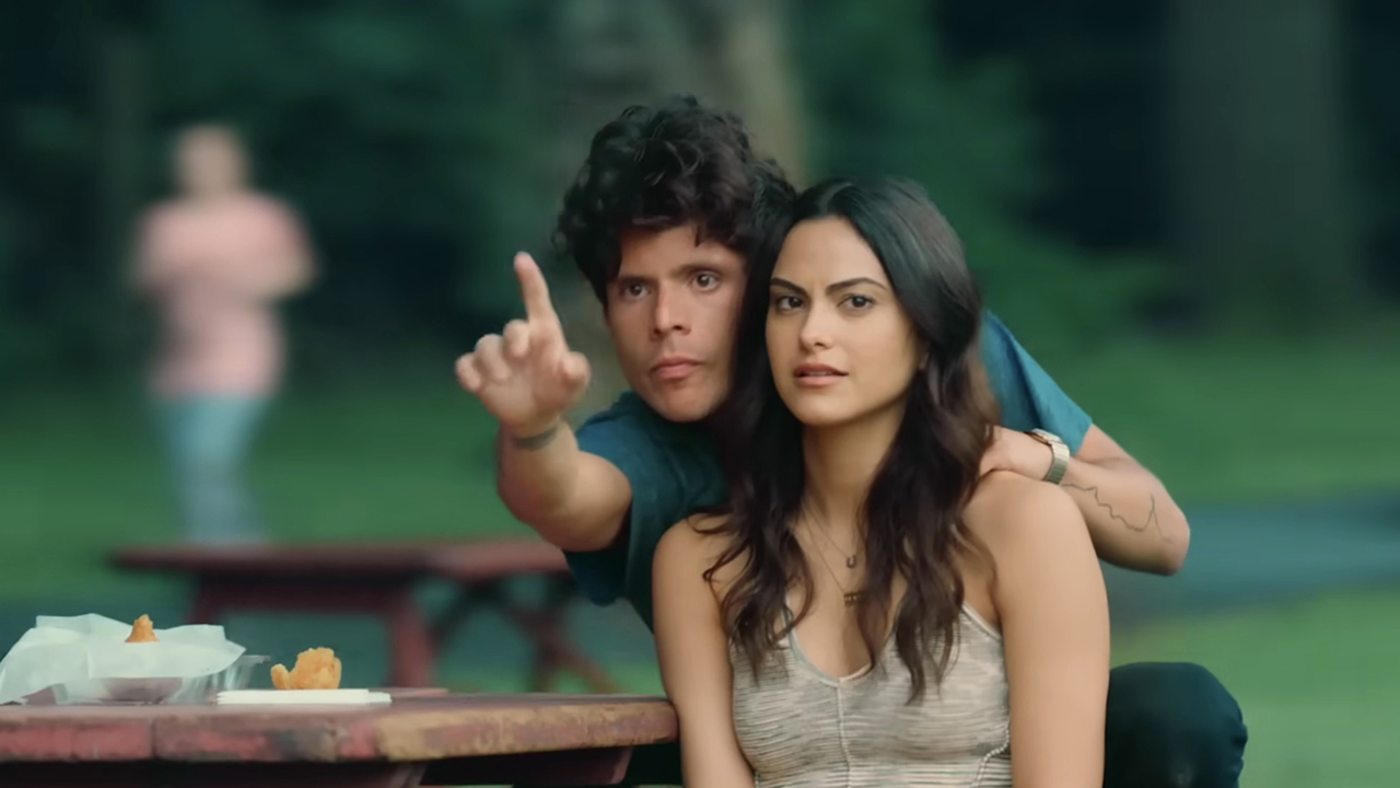 A woman and man sitting on a bench, he behind her with one hand on her shoulder and the other pointing ahead in a scene from Musica.