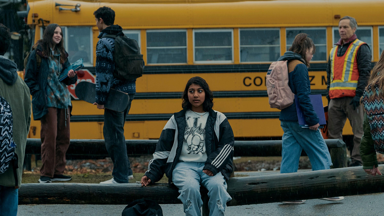 A young Indian girl sits on a bench in front of a school bus in a scene from Under the Bridge.
