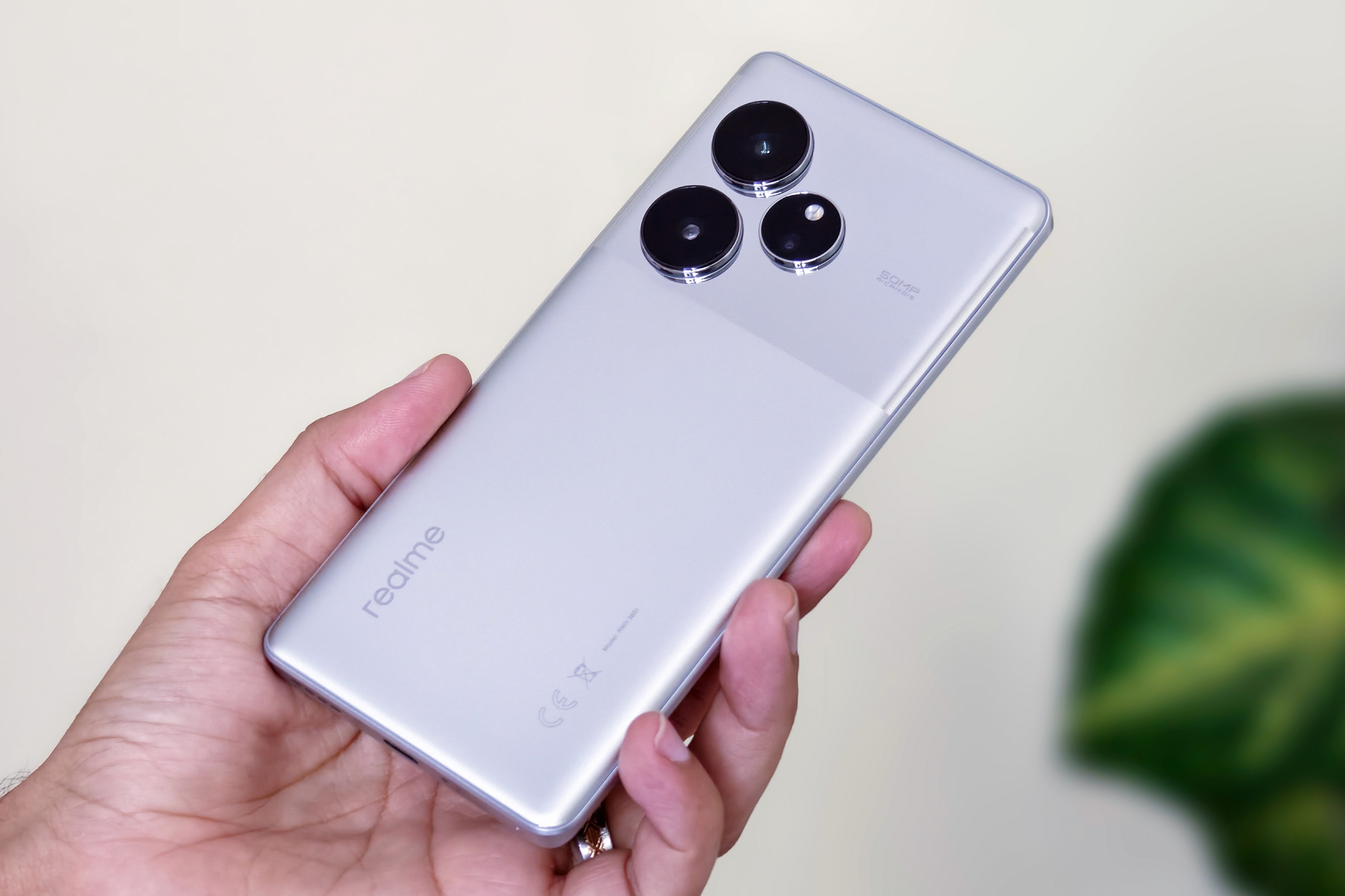 A silver Realme GT 6 Android smartphone held in hand in front of foliage and an off-white wall.